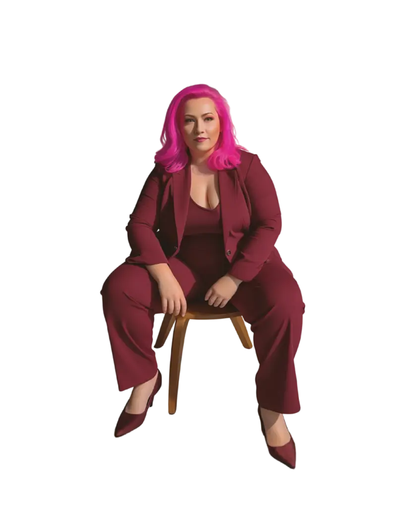 Person with bright pink hair seated on a wooden chair, wearing a burgundy suit and blazer, facing the camera.